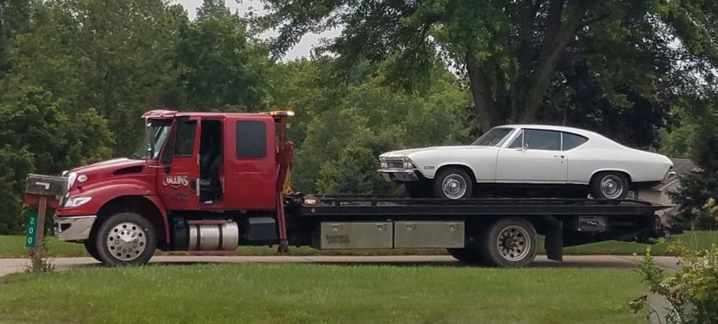 Červený ťahač vezúci biele klasické americké svalové auto po ceste.