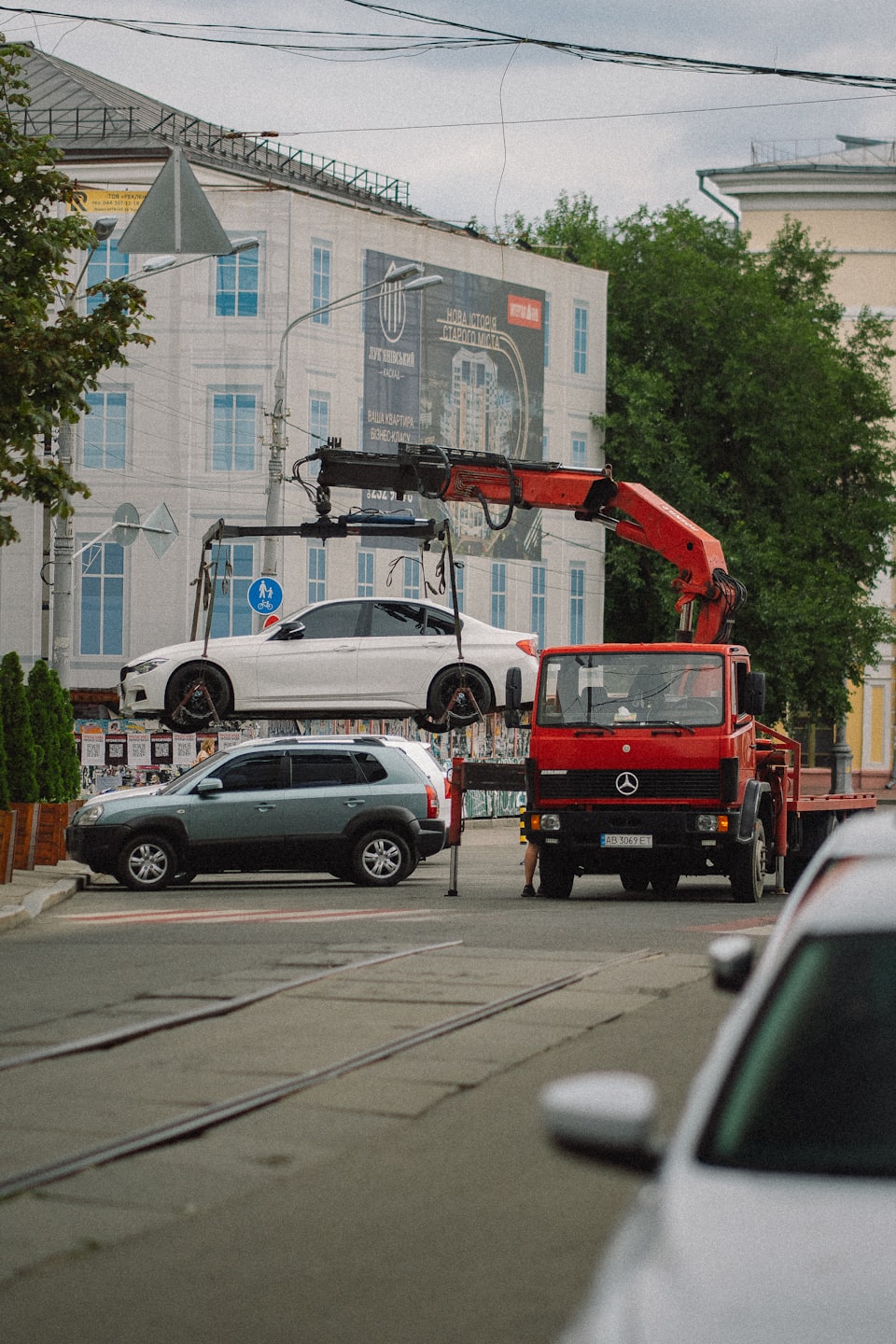 Červený odťahový vozík zdvíha biele auto na mestskej ulici.